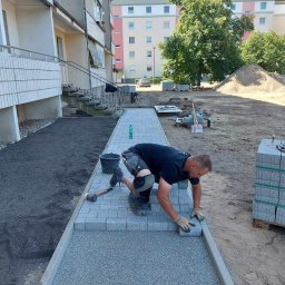 ARTELMET - Pracownik w rękawicach układa szare kostki brukowe na przygotowanym podłożu, widoczne narzędzia, budynek mieszkalny w tle.