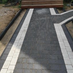 ARTELMET - Ścieżka z ciemnoszarej kostki brukowej z jasnymi pasami prowadząca do drewnianych schodów wejściowych do domu, widok z góry.