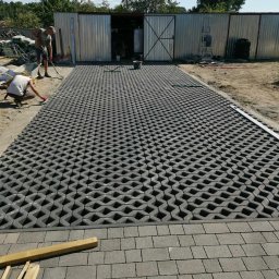 ARTELMET - Układanie nawierzchni z ażurowych płyt betonowych na podjeździe do blaszaka, dwóch robotników w trakcie pracy, widoczne narzędzia i materiały budowlane.