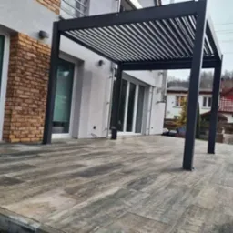 Nowoczesna, czarna pergola z lamelami na tarasie wyłożonym płytami imitującymi drewno, przylegająca do budynku z elementami cegły na elewacji.