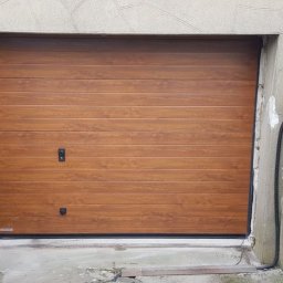 ARTELMET - Zewnętrzna, segmentowa brama garażowa w kolorze jasnego drewna, z czarnymi uchwytami i uszczelkami, zamontowana w betonowym otworze w starym budynku.