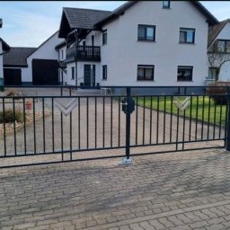 ARTELMET - Czarna, metalowa brama dwuskrzydłowa z dekoracyjnymi elementami w kształcie strzałek, zamontowana na posesji z kostki brukowej przed białymi domami.