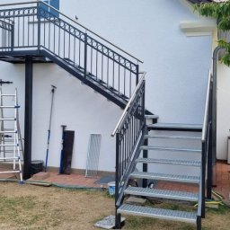 ARTELMET - Zewnętrzne schody stalowe z balustradą, prowadzące na balkon z kutą balustradą przy białym budynku, widoczne narzędzia pracy i drabina.