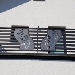 ARTELMET - Czarna, pozioma balustrada balkonowa z dekoracyjnymi, wycinanymi laserowo panelami z motywem kwiatowym na tle białej elewacji domu.