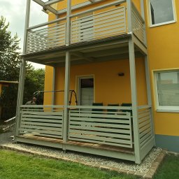 ARTELMET - Żółty dom z dwupoziomowym balkonem z szarymi, poziomymi balustradami, widok na ogród i częściowo widocznego pracownika z drabinką.