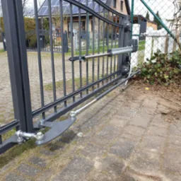 Szara brama wjazdowa z automatyką, widoczne elementy mocujące i siłownik, w tle dom z panelami słonecznymi.