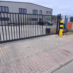 ARTELMET - Czarna, przesuwna brama wjazdowa z pionowymi prętami, widoczna z zewnątrz, z żółto-czarnym słupkiem ostrzegawczym po prawej stronie, na tle szarego budynku przemysłowego i dwóch samochodów osobowych.