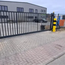 Czarna, przesuwna brama wjazdowa z pionowymi prętami, widoczna z zewnątrz, z żółto-czarnym słupkiem ostrzegawczym po prawej stronie, na tle szarego budynku przemysłowego i dwóch samochodów osobowych.