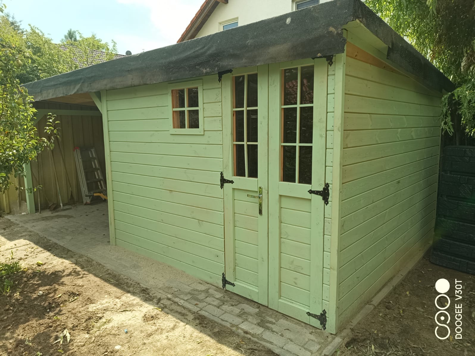 Świeżo pomalowany na pastelowy zielony kolor drewniany domek narzędziowy z dwuskrzydłowymi drzwiami z szybkami i małym okienkiem, czarne zawiasy i klamki, zadaszenie pokryte papą, widoczne...