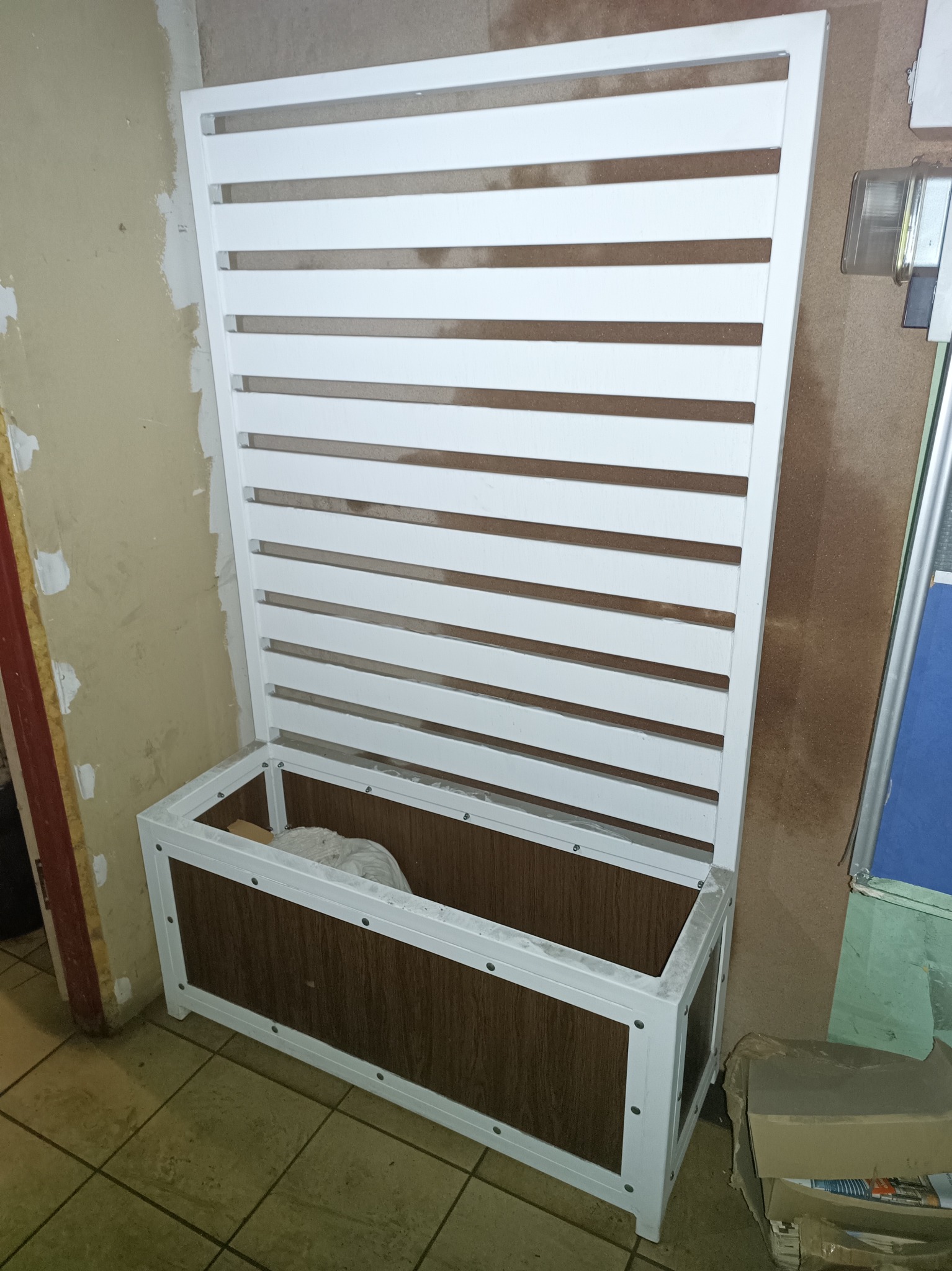 Konstrukcja z białych, poziomych listew z drewnianą skrzynią u dołu, prawdopodobnie element zabudowy lub mebel w trakcie montażu.