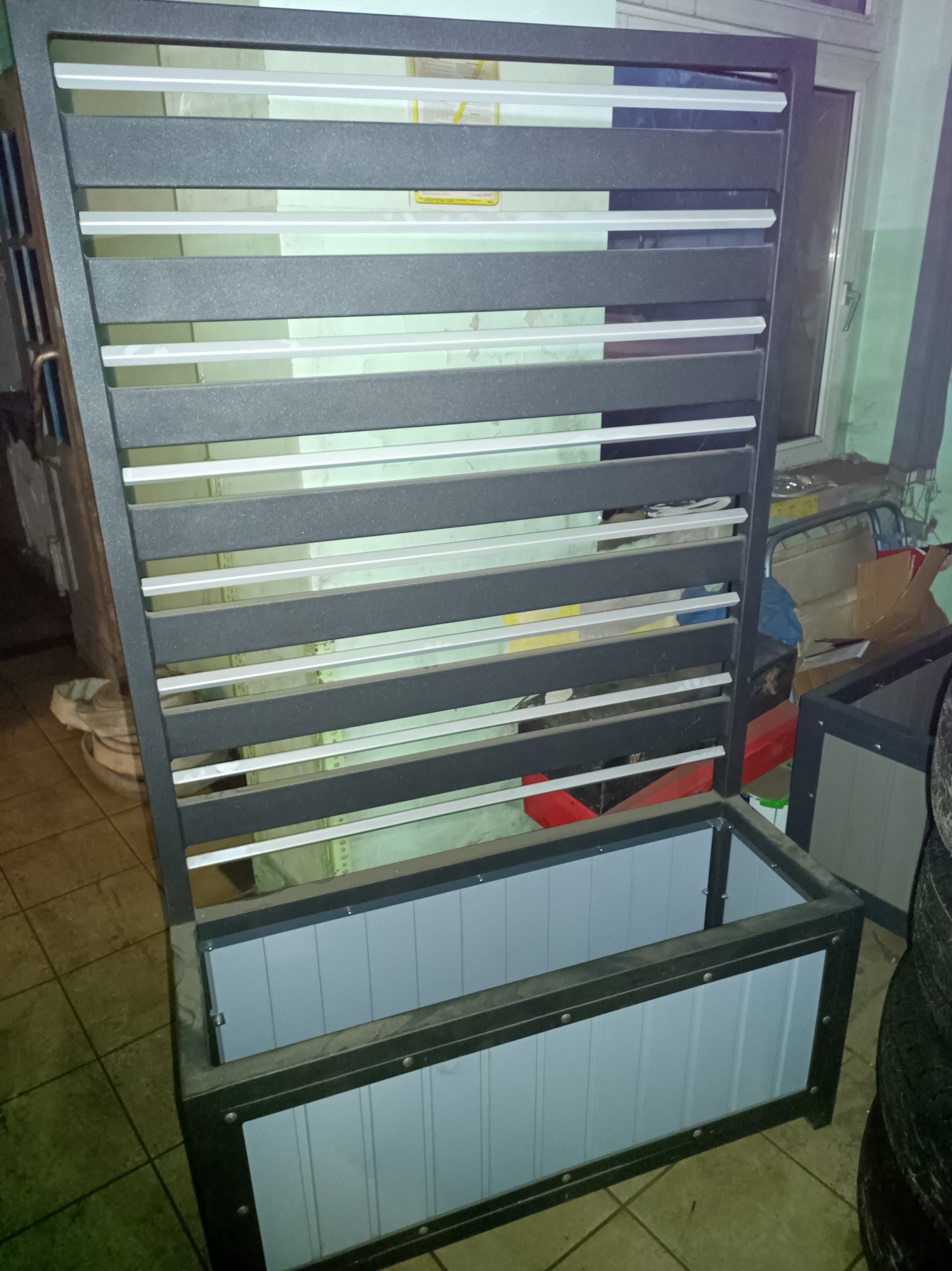 Metalowa konstrukcja z poziomymi lamelami i dolną skrzynią, prawdopodobnie element ogrodzenia lub dekoracji, w trakcie montażu lub przygotowania, widoczny w warsztacie.