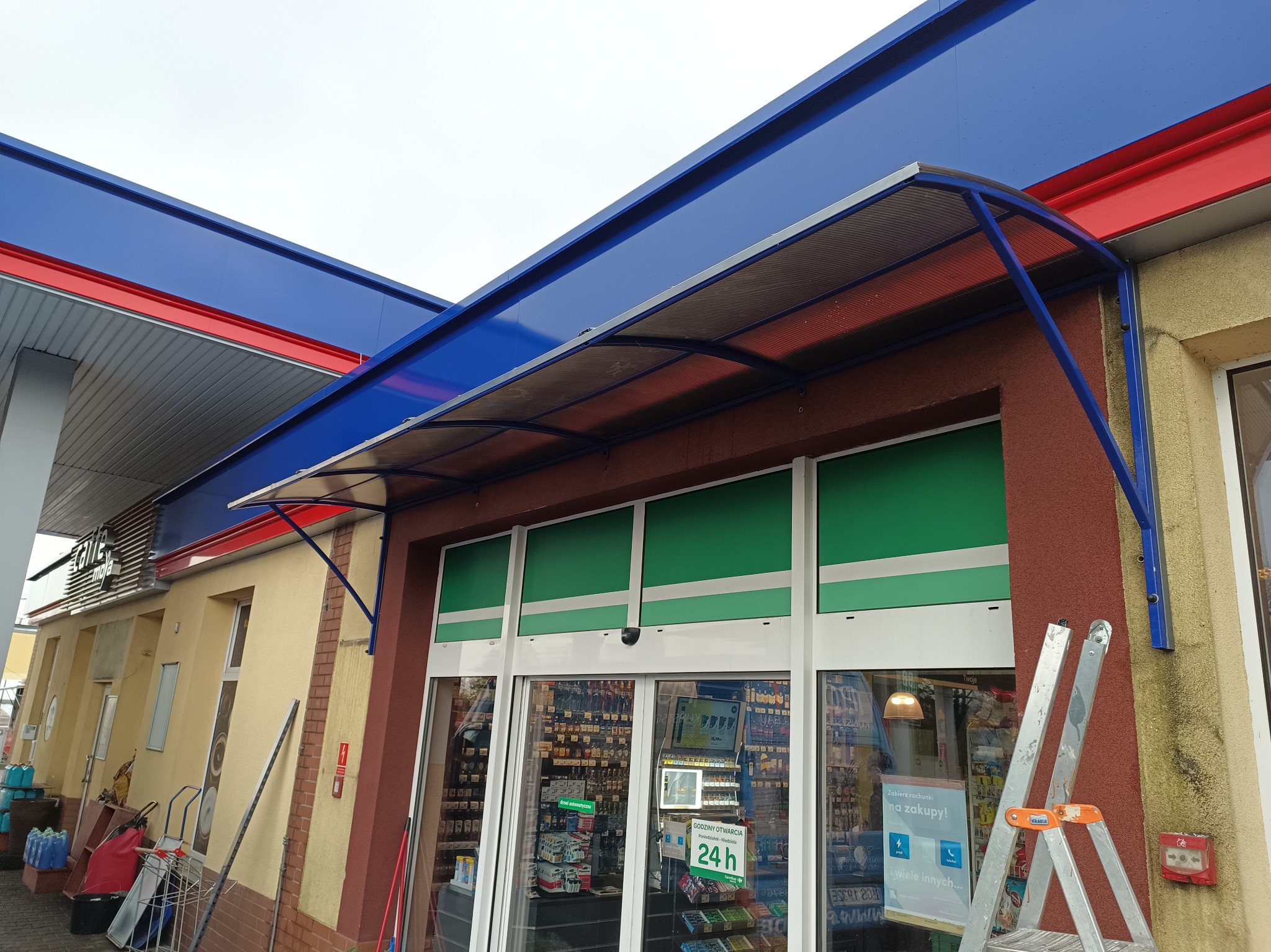 Zadaszenie nad wejściem do sklepu na stacji benzynowej, konstrukcja z niebieskich profili i przezroczystego poliwęglanu, widoczna drabina oparta o ścianę.