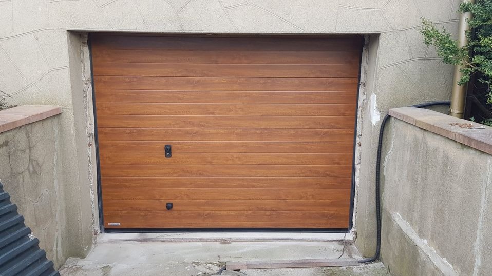 Zewnętrzna, segmentowa brama garażowa w kolorze jasnego drewna, z czarnymi uchwytami i uszczelkami, zamontowana w betonowym otworze w starym budynku.