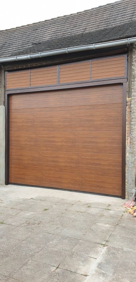 Zewnętrzna brama garażowa segmentowa w kolorze drewna z poziomymi panelami, zamontowana w budynku z szarą dachówką i elementami ceglanymi.