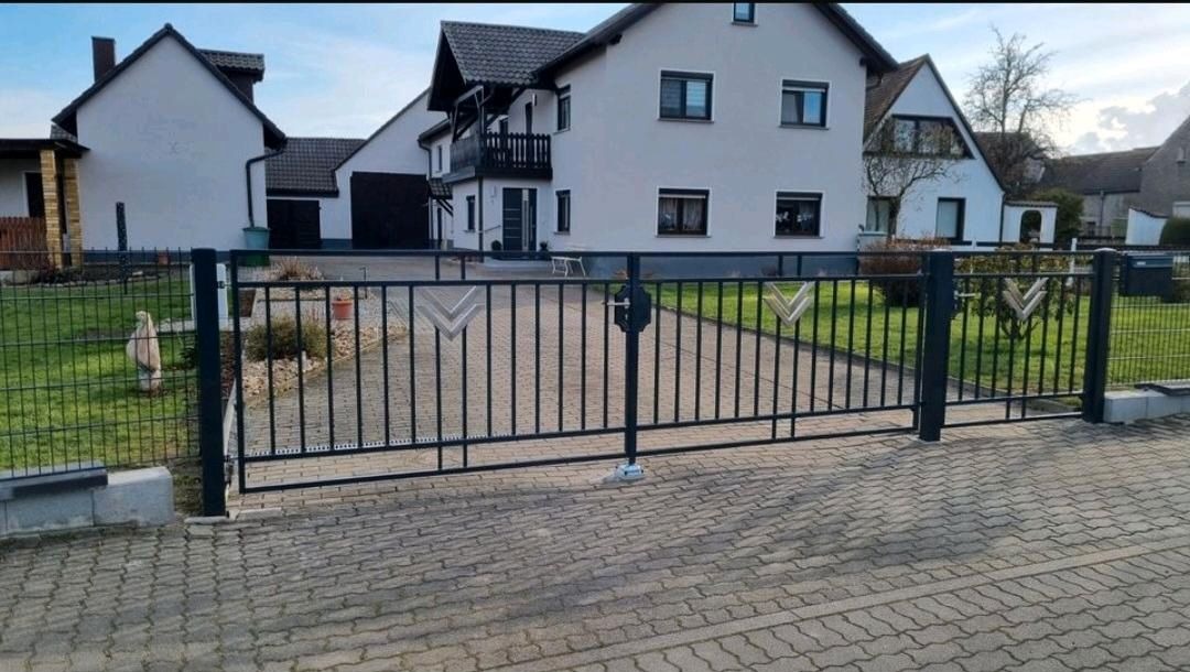 Czarna, metalowa brama dwuskrzydłowa z dekoracyjnymi elementami w kształcie strzałek, zamontowana na posesji z kostki brukowej przed białymi domami.