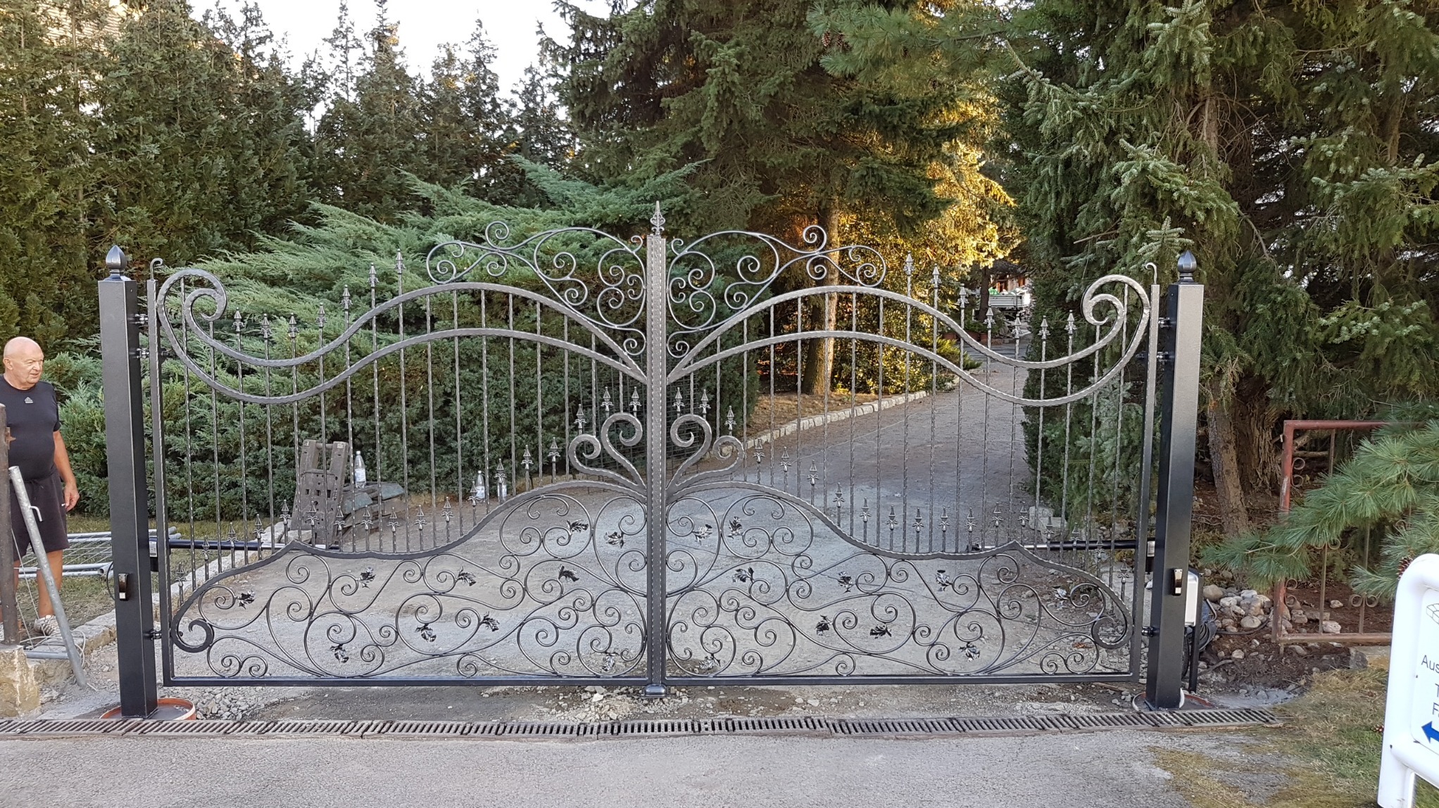 Otwarta, metalowa brama wjazdowa z ozdobnymi, kutymi elementami na tle zielonych drzew i krzewów; widoczny fragment napędu i mężczyzna w czarnej koszulce.
