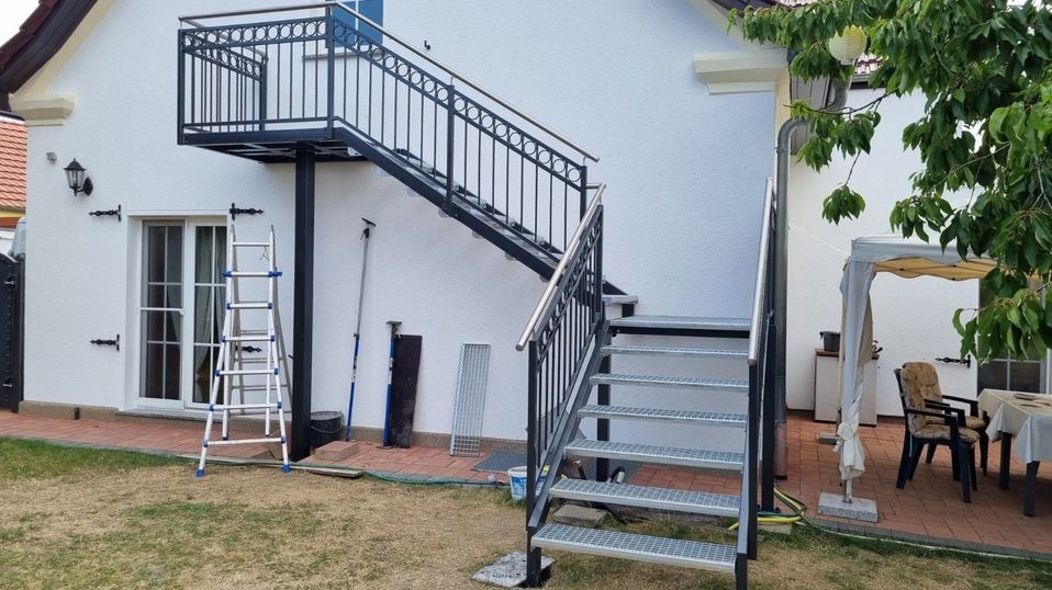 Zewnętrzne schody stalowe z balustradą, prowadzące na balkon z kutą balustradą przy białym budynku, widoczne narzędzia pracy i drabina.