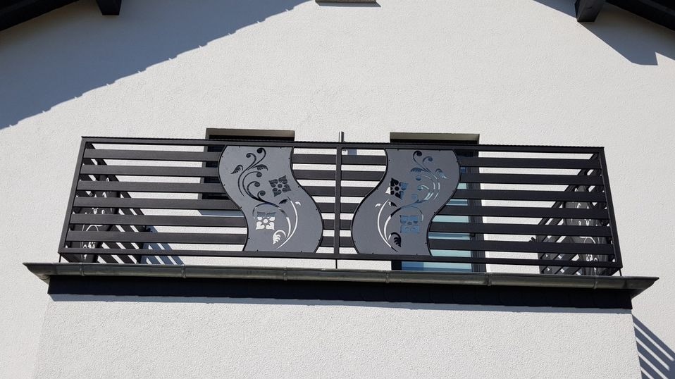 Czarna, pozioma balustrada balkonowa z dekoracyjnymi, wycinanymi laserowo panelami z motywem kwiatowym na tle białej elewacji domu.