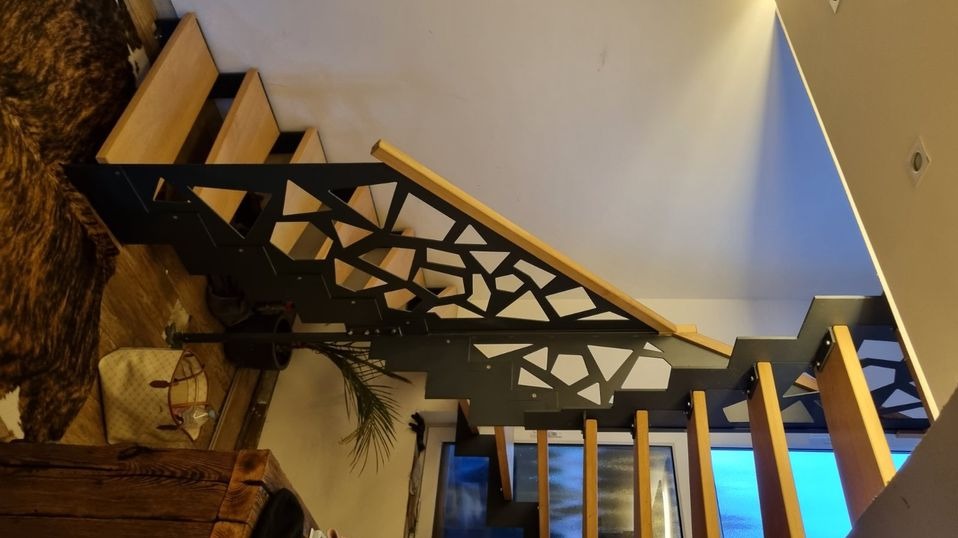 Nowoczesne schody drewniane z czarną, metalową balustradą o geometrycznym wzorze, widok z dołu.