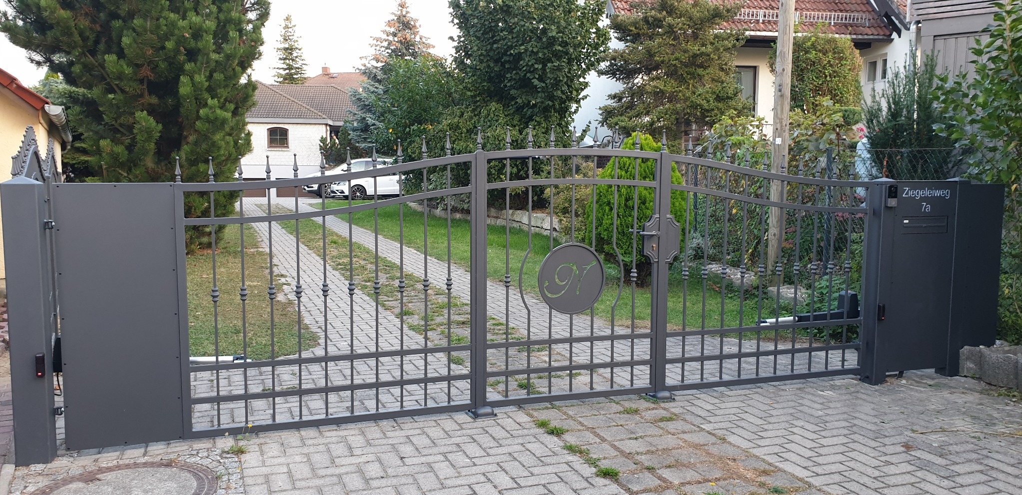 Szara, metalowa brama wjazdowa z dekoracyjnym monogramem na tle brukowanej drogi dojazdowej i domu.