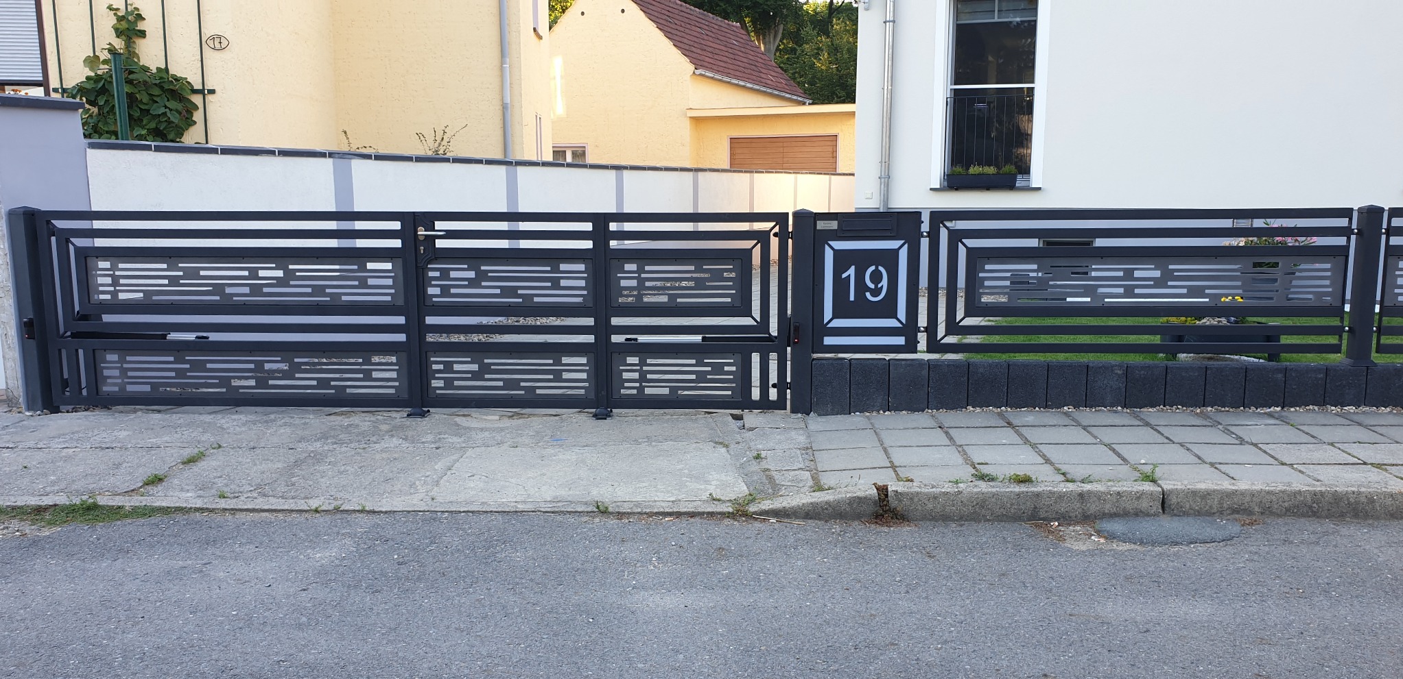 Nowoczesne, metalowe ogrodzenie posesji z bramą wjazdową o geometrycznym wzorze, z numerem domu '19' na słupku.