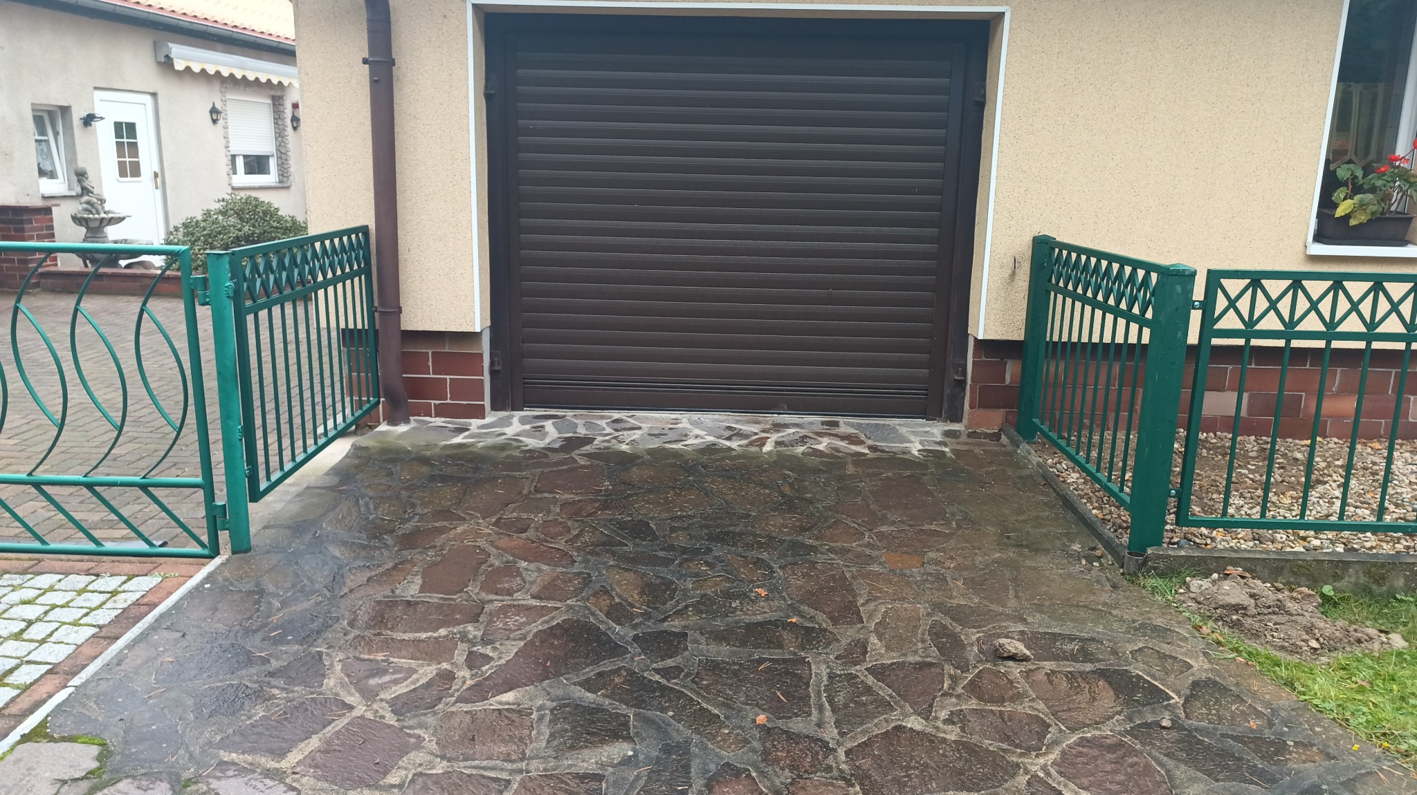 Brązowa brama rolowana garażowa, zielone metalowe ogrodzenie z dwoma skrzydłami bramki, kamienny podjazd na posesji.