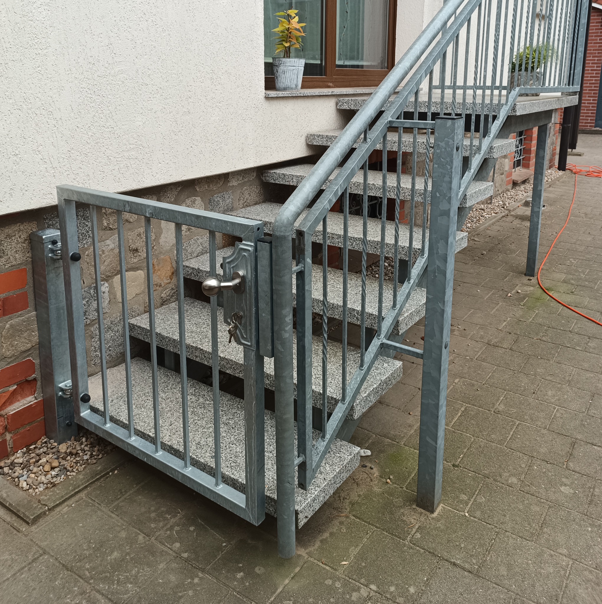 Metalowa furtka z zamkiem przy schodach zewnętrznych z szarego lastryko, widoczne klucze w zamku i doniczka z rośliną na parapecie.