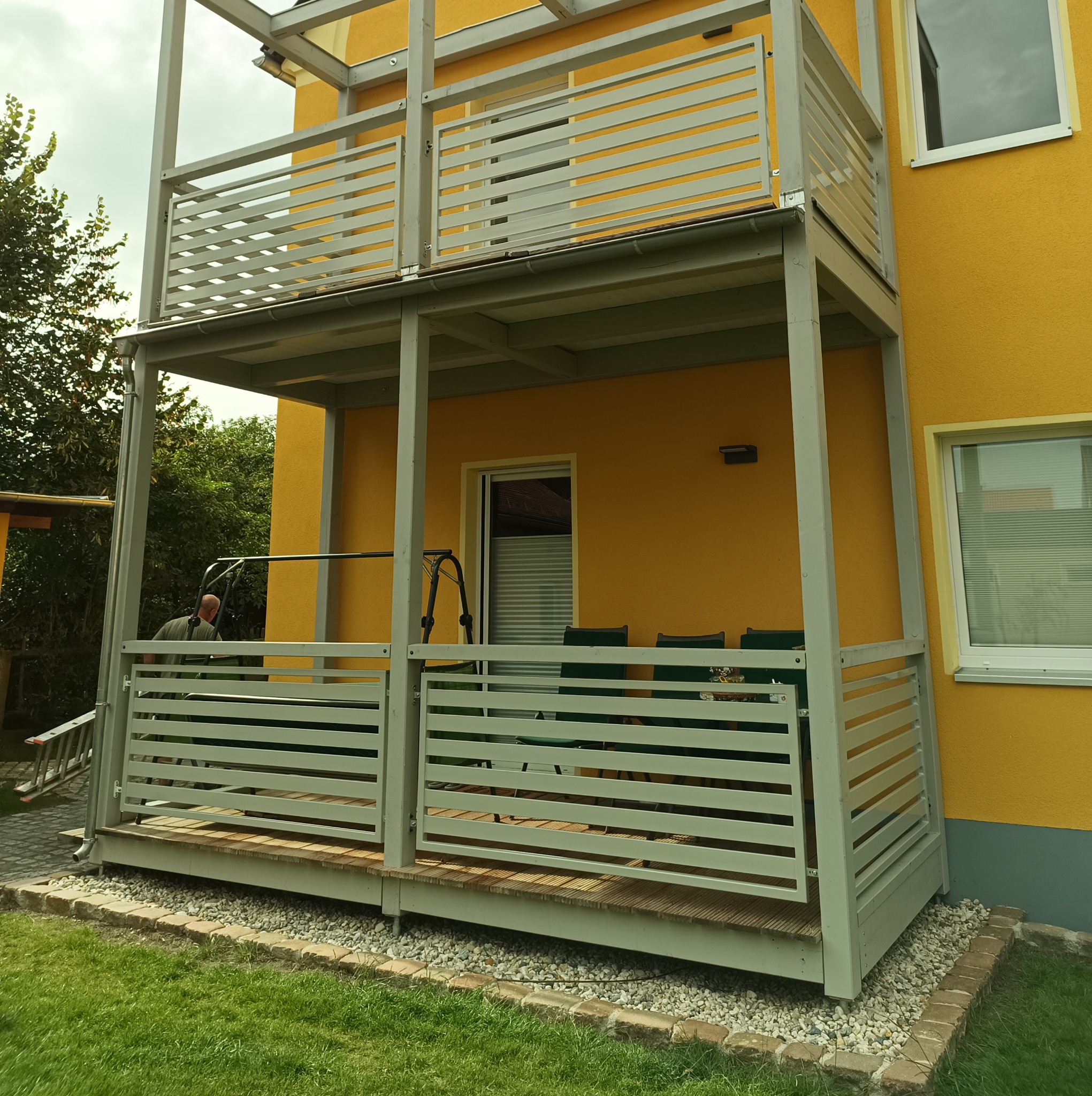 Żółty dom z dwupoziomowym balkonem z szarymi, poziomymi balustradami, widok na ogród i częściowo widocznego pracownika z drabinką.