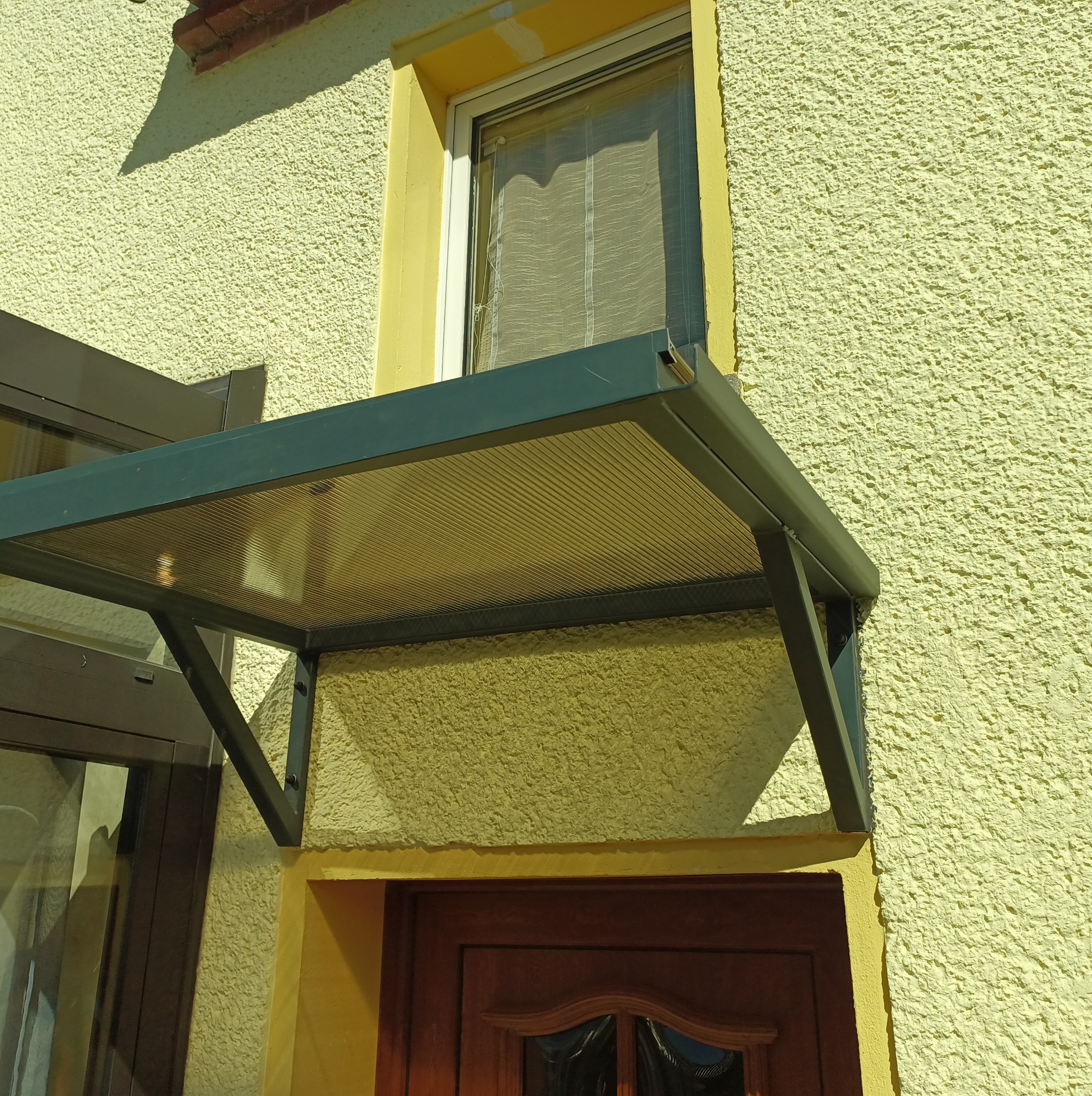 Zadaszenie nad drzwiami wejściowymi z przezroczystym panelem i metalową konstrukcją, zamontowane na elewacji budynku o żółtym, ziarnistym tynku.