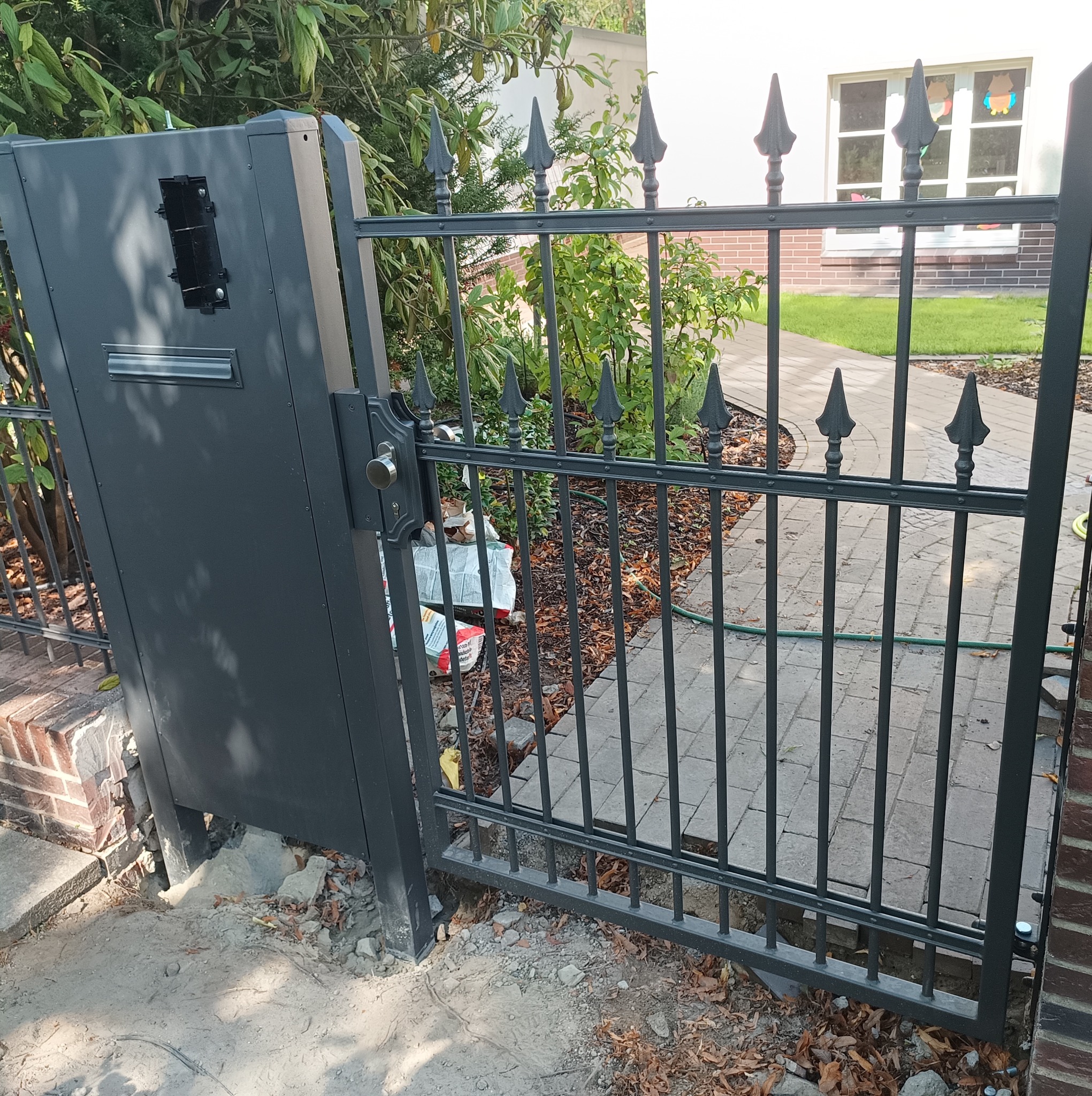 Otwarta, grafitowa brama wjazdowa z dekoracyjnymi grotami, widoczne ślady montażu i worki z zaprawą w tle.