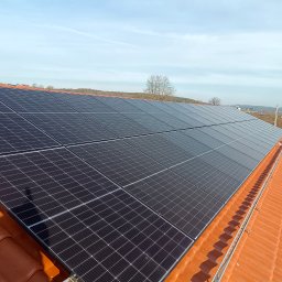 USŁUGI REMONTOWO-BUDOWLANE DANIEL MOSKALEK - Panele fotowoltaiczne zamontowane na pomarańczowej dachówce, widok z góry na tle krajobrazu z drzewami i budynkami.