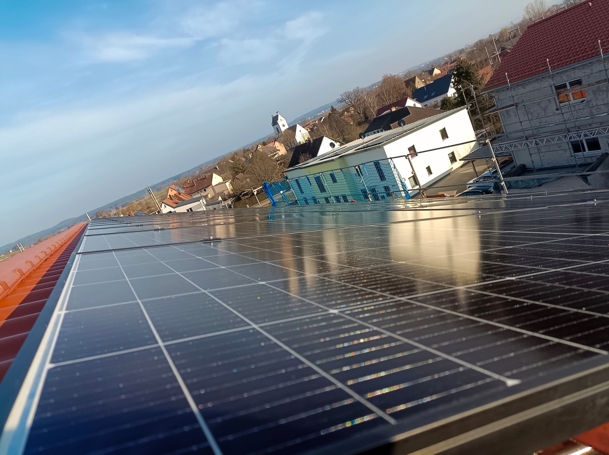 Widok z góry na panele fotowoltaiczne zamontowane na czerwonym dachu budynku, z odbiciem błękitnego nieba i zabudowań w tle.