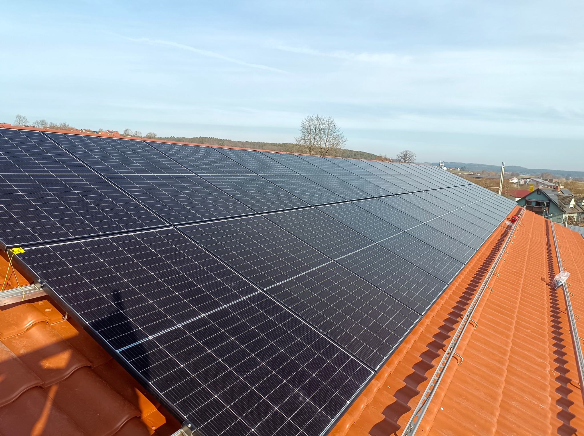 Panele fotowoltaiczne zamontowane na pomarańczowej dachówce, widok z góry na tle krajobrazu z drzewami i budynkami.