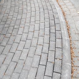 VERSA CORP SPÓŁKA Z OGRANICZONĄ ODPOWIEDZIALNOŚCIĄ - Szara kostka brukowa ułożona łukiem, oddzielona obrzeżem od asfaltu. Widoczne drobne liście na powierzchni. Ujęcie z góry, podkreślające wzór ułożenia.