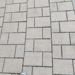 VERSA CORP SPÓŁKA Z OGRANICZONĄ ODPOWIEDZIALNOŚCIĄ - Szara kostka brukowa ułożona w regularny wzór, widoczne buty robocze w dolnej części kadru. Układanie w prostokąty, widok z góry.