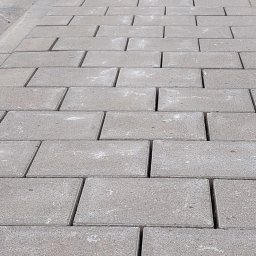 VERSA CORP SPÓŁKA Z OGRANICZONĄ ODPOWIEDZIALNOŚCIĄ - Szara kostka brukowa ułożona w regularny wzór, widok z perspektywy, z delikatnym cieniem podkreślającym fakturę. Minimalistyczny design chodnika.