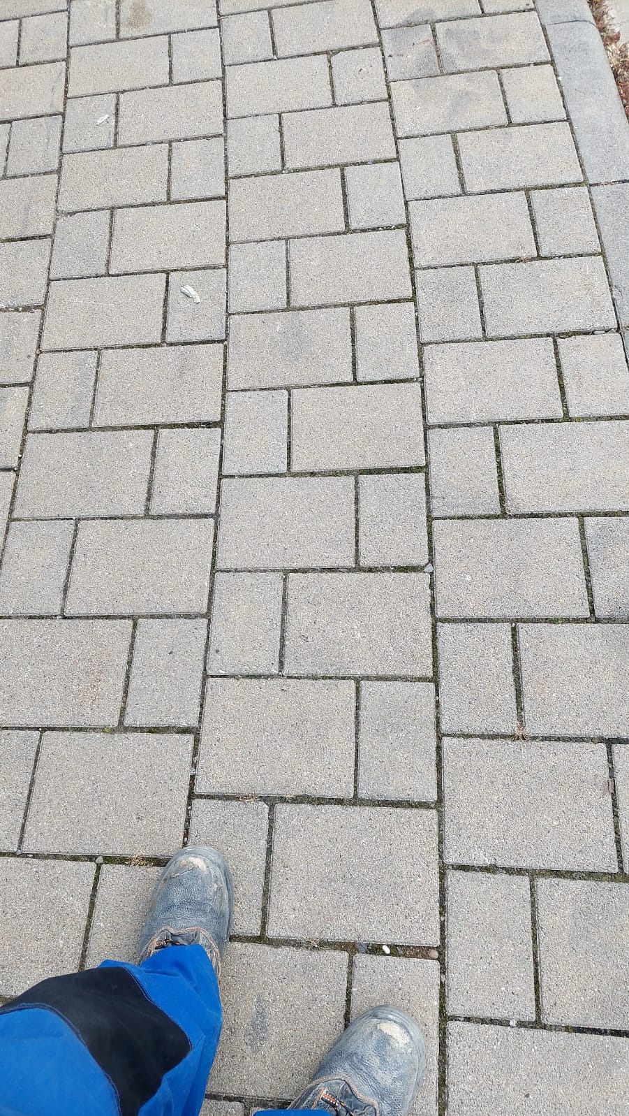 Szara kostka brukowa ułożona w regularny wzór, widoczne buty robocze w dolnej części kadru. Układanie w prostokąty, widok z góry.