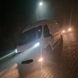 Biały bus dostawczy oświetlony reflektorami na brukowanej drodze nocą, widoczny pod lampą uliczną i w oddali światła innego pojazdu.