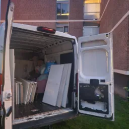 Otwarty bagażnik białego busa załadowany meblami, kartonami i torbami podczas przeprowadzki, na tle ceglanego budynku mieszkalnego i trawnika.