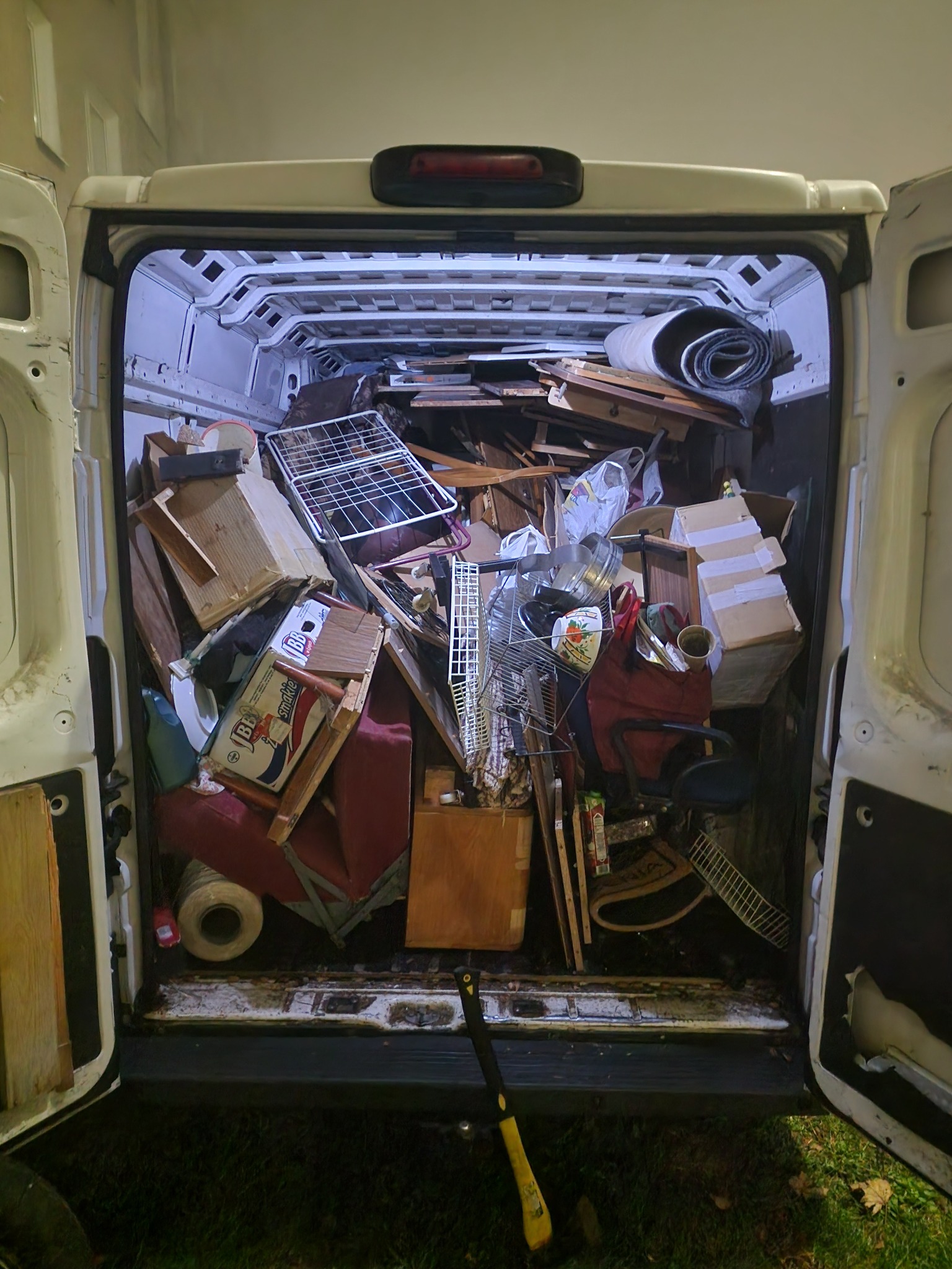 Wnętrze białego furgonetki załadowane różnorodnymi przedmiotami: meblami, kartonami, sprzętem AGD, elementami wyposażenia wnętrz, widoczne otwarte tylne drzwi pojazdu i fragment trawnika z żółtą...