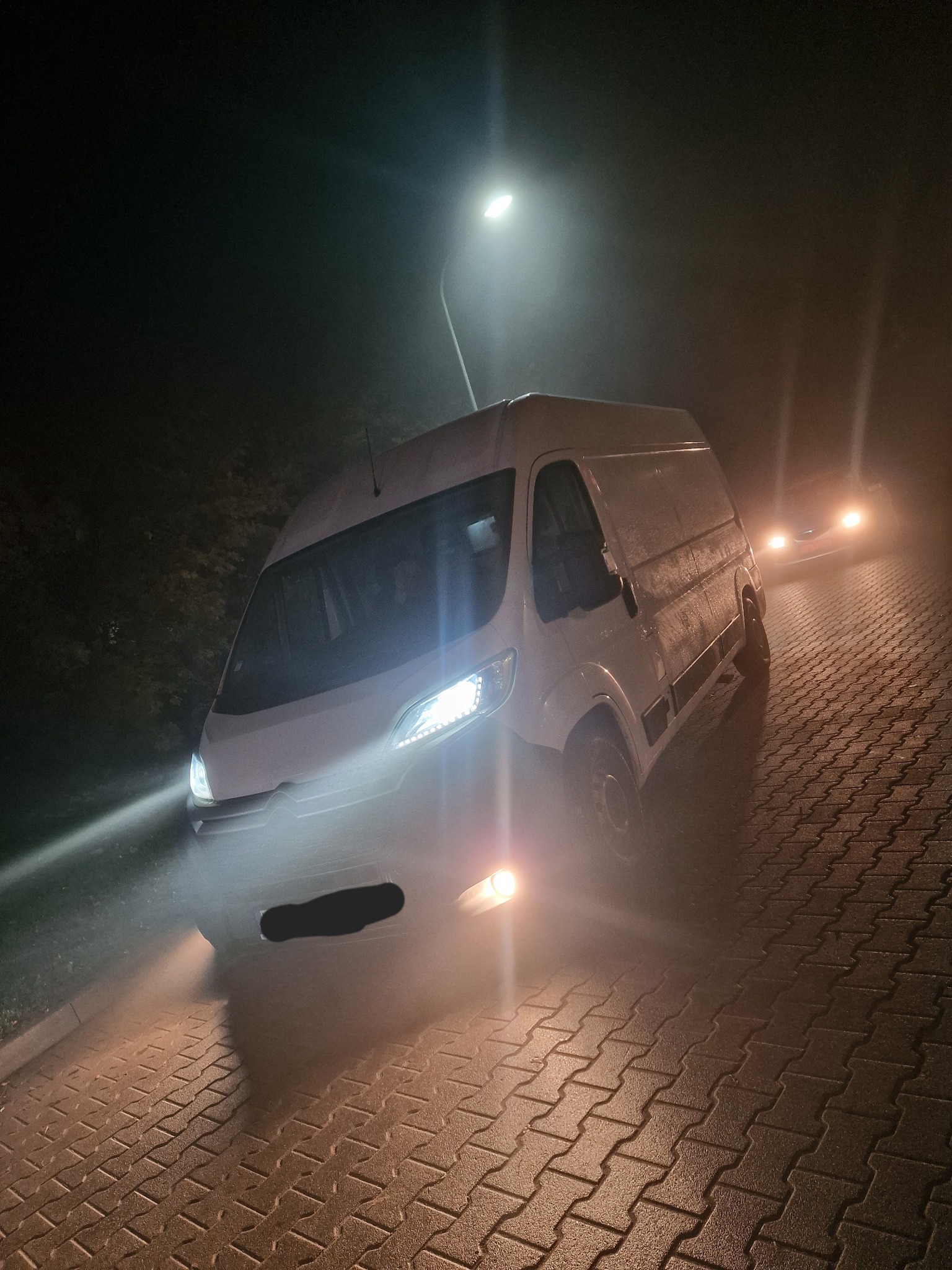 Biały bus dostawczy oświetlony reflektorami na brukowanej drodze nocą, widoczny pod lampą uliczną i w oddali światła innego pojazdu.
