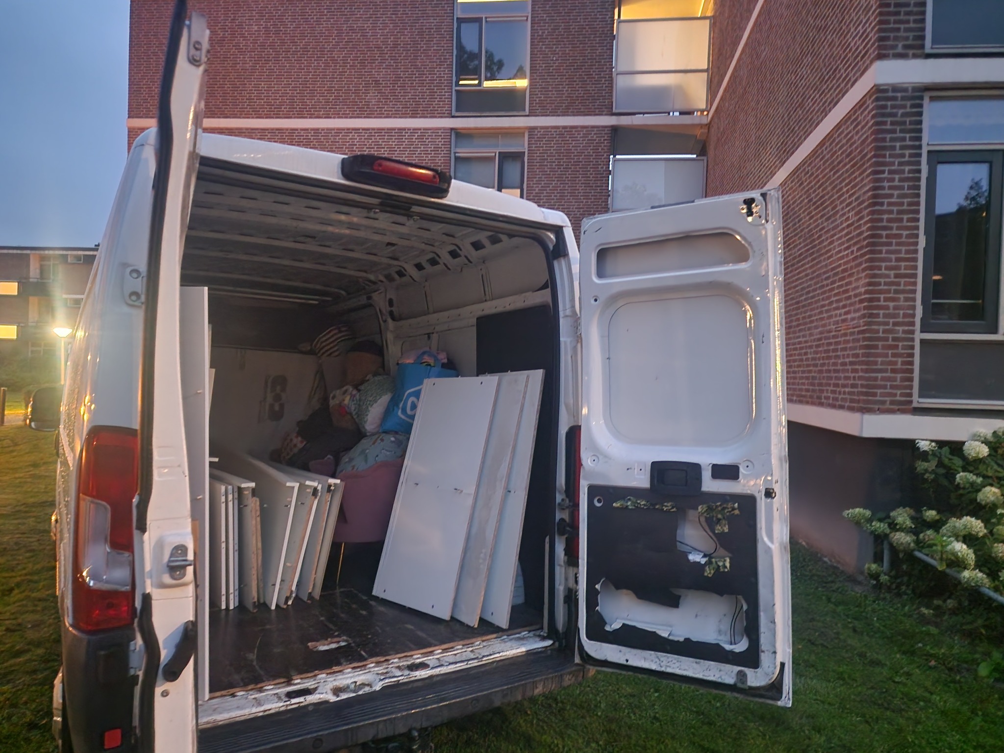 Otwarty bagażnik białego busa załadowany meblami, kartonami i torbami podczas przeprowadzki, na tle ceglanego budynku mieszkalnego i trawnika.
