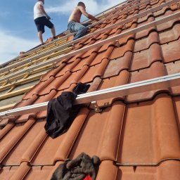 BUD-IK Ivan Kok - Ekipa dekarzy montuje aluminiowe profile na dachu z czerwonej dachówki ceramicznej, widoczne fragmenty drewnianej konstrukcji, w kadrze rękawice robocze i czarna bluza.