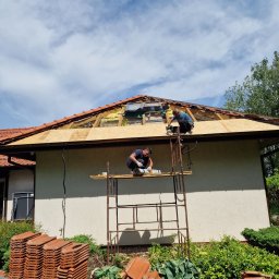 BUD-IK Ivan Kok - Dwóch dekarzy na rusztowaniu montuje nowe poszycie dachu z płyt OSB, widoczne stosy dachówek ceramicznych na paletach przed budynkiem.
