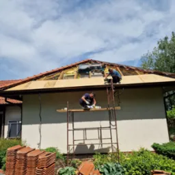 Dwóch dekarzy na rusztowaniu montuje nowe poszycie dachu z płyt OSB, widoczne stosy dachówek ceramicznych na paletach przed budynkiem.