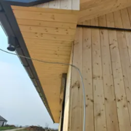 Drewniana elewacja budynku w trakcie budowy z widocznymi elementami dachu i rynną, na tle krajobrazu z domem i stertą ziemi.