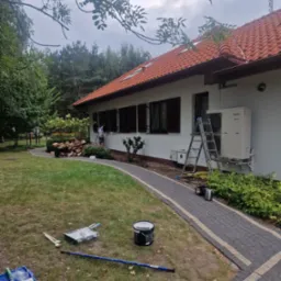 Malowanie elewacji domu jednorodzinnego z brązowymi okiennicami, widoczne narzędzia malarskie rozłożone na trawniku, drabina oparta o ścianę, w tle wózek z drewnem.