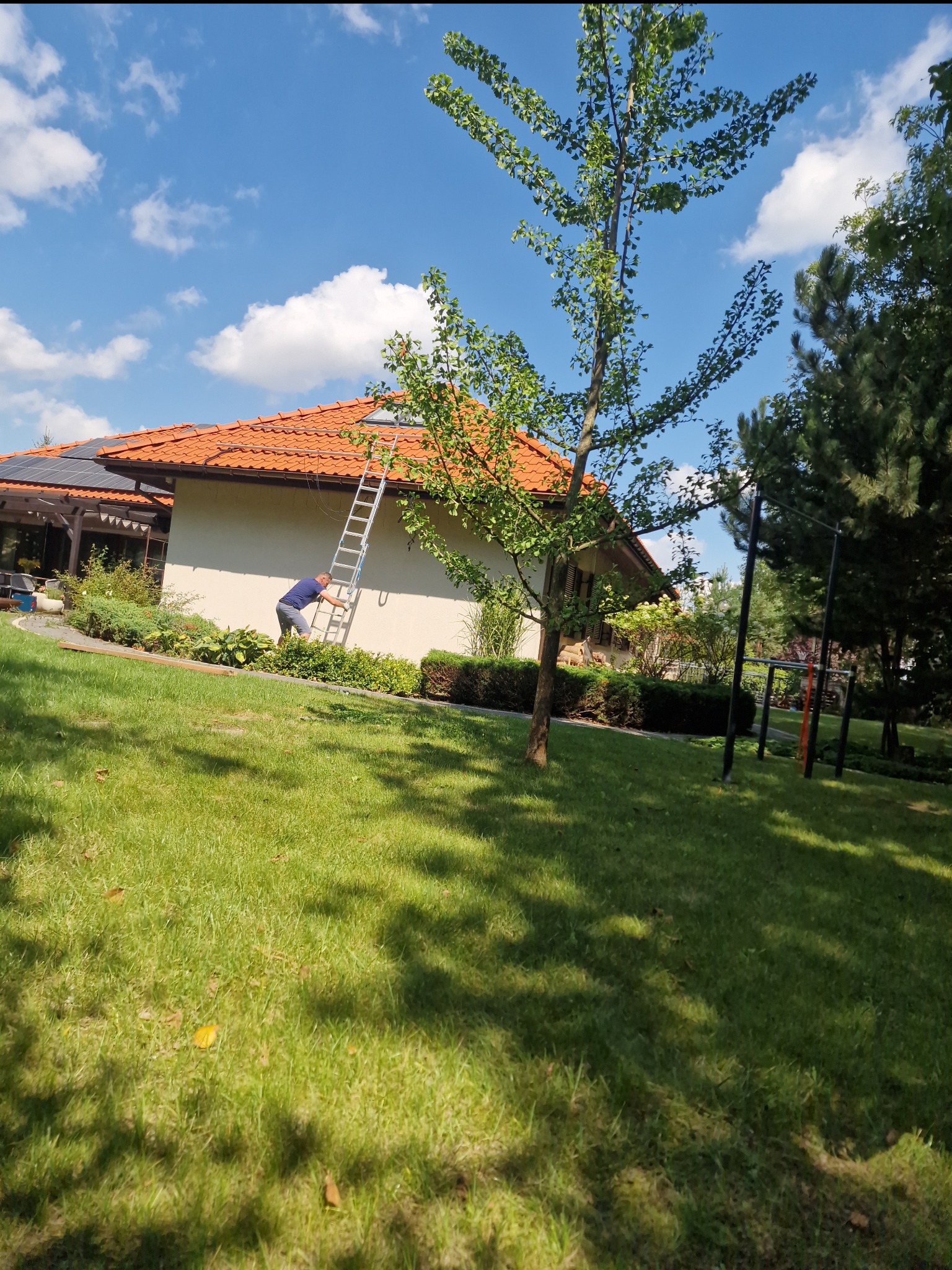 Pracownik na drabinie przy ścianie domu z pomarańczową dachówką, widoczne panele słoneczne, trawnik i drzewa w słoneczny dzień.