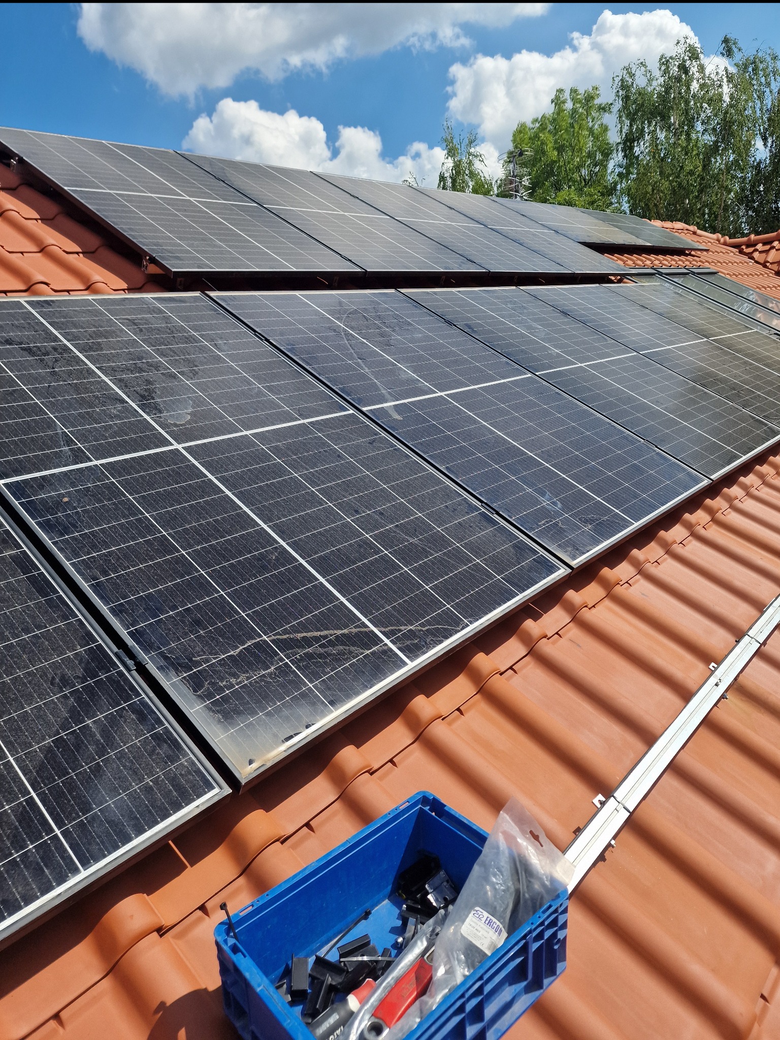 Montaż paneli fotowoltaicznych na dachu z czerwonej dachówki, widoczna skrzynka z narzędziami i elementami montażowymi oraz częściowo zamontowana szyna montażowa.