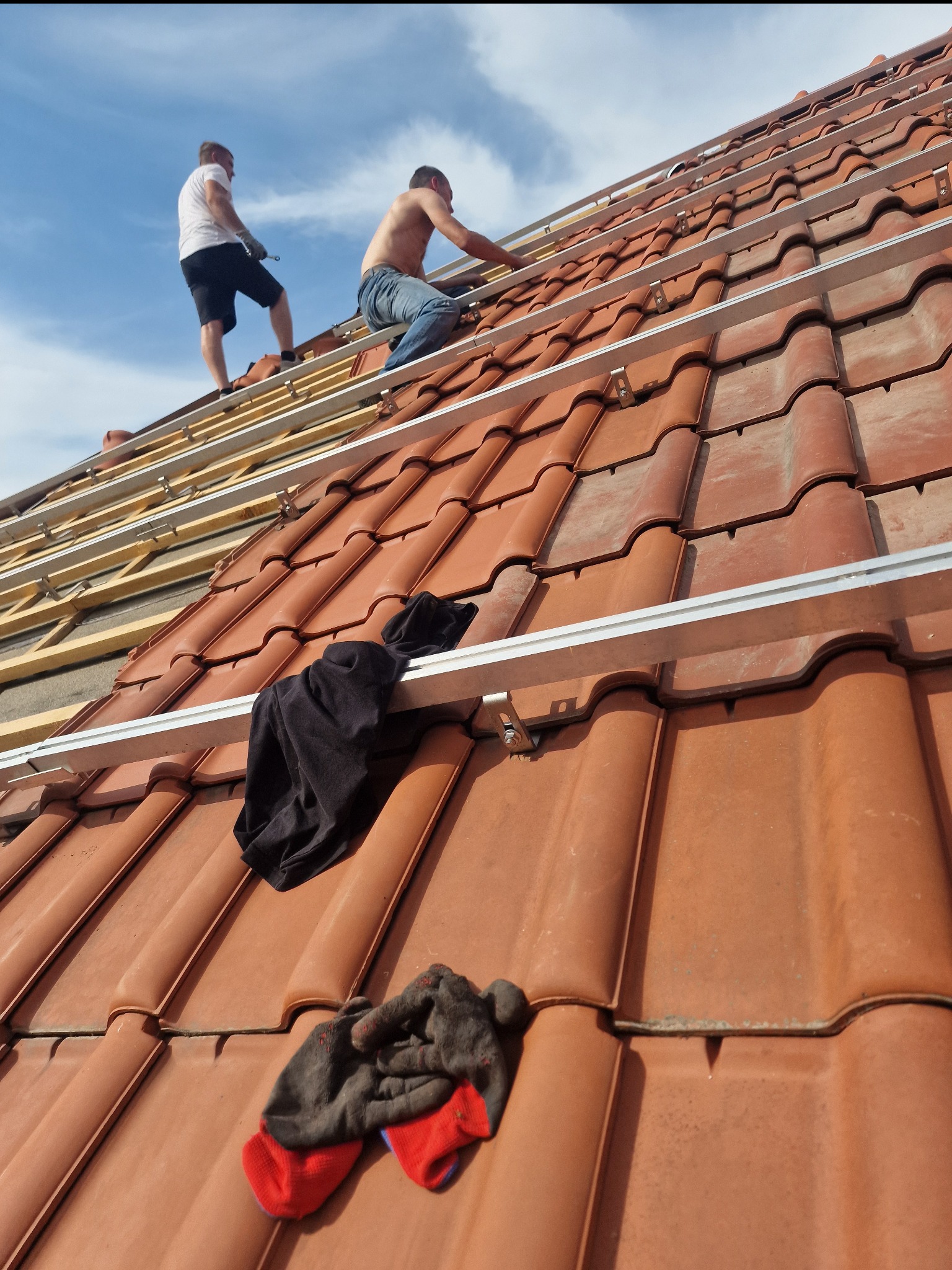 Ekipa dekarzy montuje aluminiowe profile na dachu z czerwonej dachówki ceramicznej, widoczne fragmenty drewnianej konstrukcji, w kadrze rękawice robocze i czarna bluza.